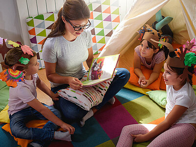 Eine Erwachsene liest in einem bunten Zelt aus einem Buch drei Kindern vor, die auf farbigen Kissen sitzen und fantasievolle Kopfbedeckungen aus Papier tragen.