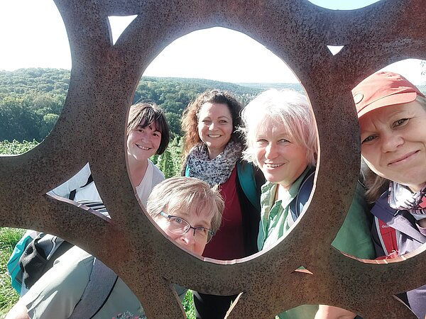 Fünf Frauen lächeln für ein gemeinsames Foto durch die runden Aussparungen einer großen Metallskulptur. Im Hintergrund ist eine grüne, hügelige Landschaft zu sehen.