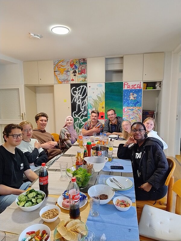 Mehrere Auszubildende sitzen um einen rechteckigen Esstisch in einem hellen Raum. Auf dem Tisch stehen verschiedene Schüsseln und Teller mit Speisen wie Gurkenscheiben, Chips, Brot, Salat und Dips. Es sind auch Getränkeflaschen, darunter eine Colaflasche, zu sehen. Im Hintergrund hängen bunte, teils handgemalte Bilder und ein Schild mit dem Namen 'Pascal' an einem Schrank. Die Personen blicken in Richtung Kamera, einige lächeln.