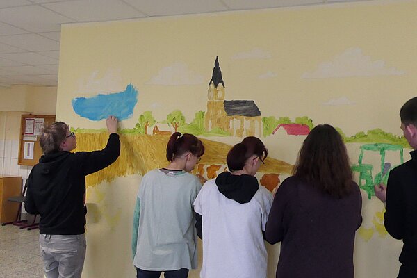 Erzieher*innen begleiten die Kreativtage an der Förderschule für geistige Entwicklung in Skäßchen Vier Personen malen eine ländliche Szene auf eine Wand. Ein Schüler malt den Himmel, während andere an den Feldern und Tieren arbeiten.