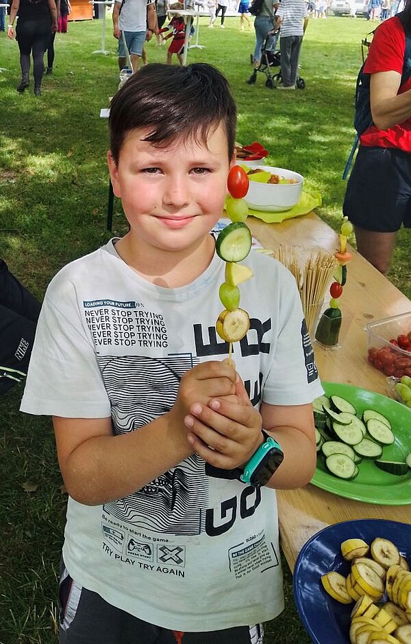 Ein lächelndes Kind hält einen selbstgemachten Obstspieß mit Trauben, Gurken, Bananen und Tomaten in den Händen. Im Hintergrund sind weitere Obstschalen und ein grüner Tisch zu sehen.