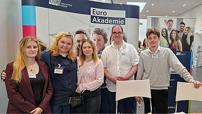 Fünf junge Erwachsene, darunter drei Frauen und zwei Männer, posieren lächelnd vor einem Messestand der Euro Akademie. Der Stand zeigt Roll-ups mit dem Logo der Euro Akademie und der ESO Education Group.