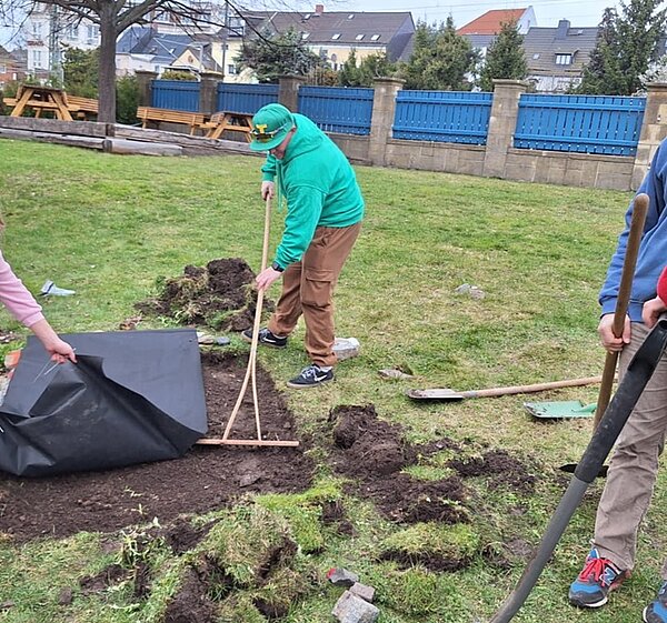 Drei Auszubildende arbeiten auf einer Rasenfläche. Eine Person hält eine Schaufel, eine andere zieht mit einem Rechen Erde glatt, und eine dritte Person hält eine schwarze Folie oder Plane. Im Hintergrund stehen Holzbänke und ein blauer Zaun, dahinter sind Wohnhäuser zu sehen. Auf dem Boden liegen Werkzeuge und ausgehobene Erde.