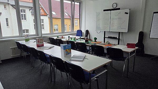 Ein Klassenzimmer mit weißen Tafeln, Tischen mit weißen Tischdecken und Büchern in Rot, Grün und Blau sowie einem roten und grünen Karton.