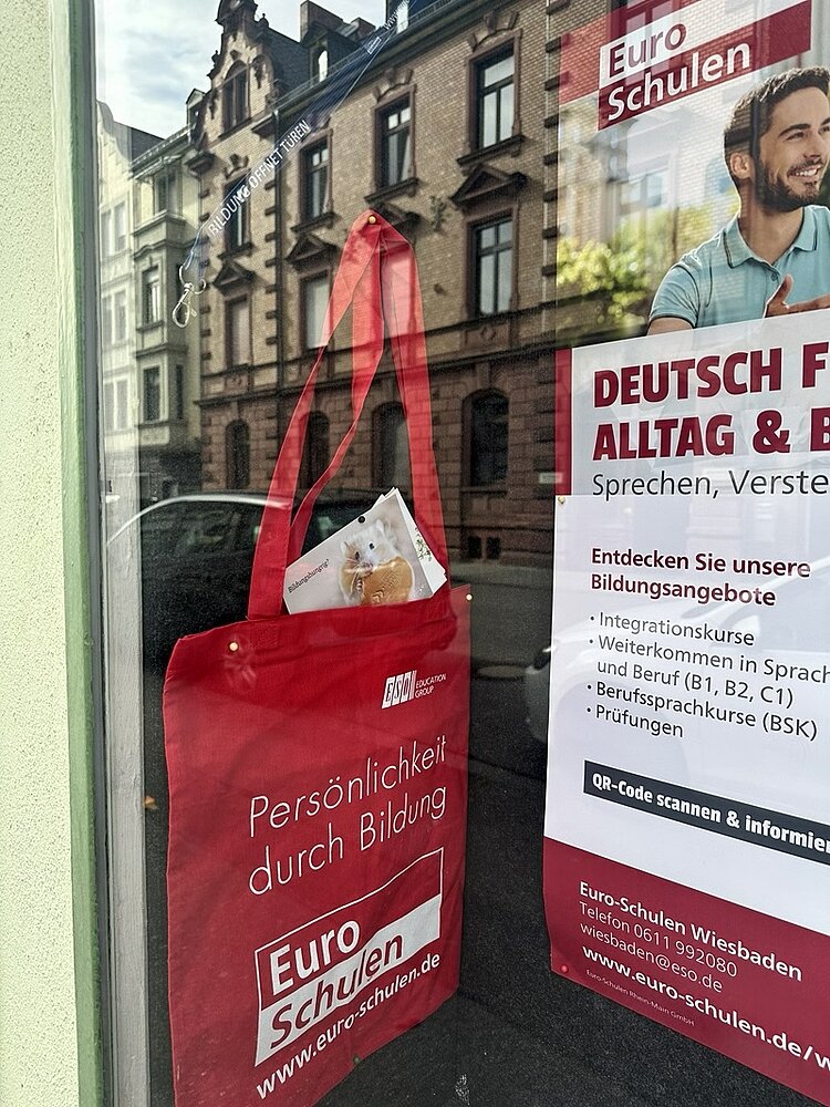 Eine rote Stofftasche mit weißem Aufdruck hängt an einem Türgriff vor einer Fensterscheibe. Auf der Tasche steht in weißer Schrift: 'Persönlichkeit durch Bildung' und darunter das Logo sowie der Name 'Euro Schulen'. Im Fenster dahinter ist ein Plakat mit weiteren Informationen zu Sprach- und Bildungsangeboten der Euro Schulen zu sehen. Die Spiegelung zeigt eine Häuserfassade mit mehreren Fenstern und Bäumen auf der gegenüberliegenden Straßenseite.