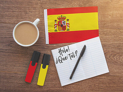 Ein Foto von einem Holztisch mit einer Tasse Kaffee, einer kleinen spanischen Flagge, zwei Textmarkern in den Farben der Flagge und einem Notizblock, auf dem 'Hola! ¿Qué tal?' geschrieben steht.