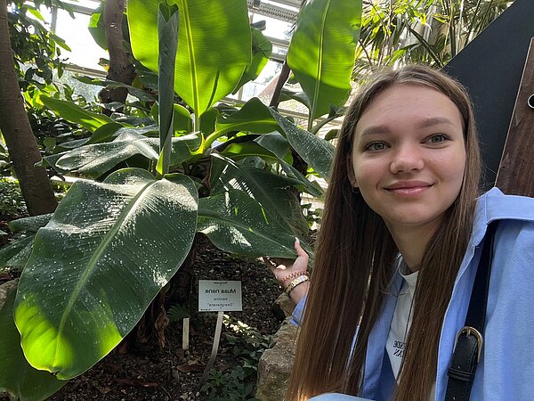 Exkursion in den Botanischen Garten Würzburg Eine junge Frau steht lächelnd neben einer großen Pflanze mit großen grünen Blättern in einem Gewächshaus.