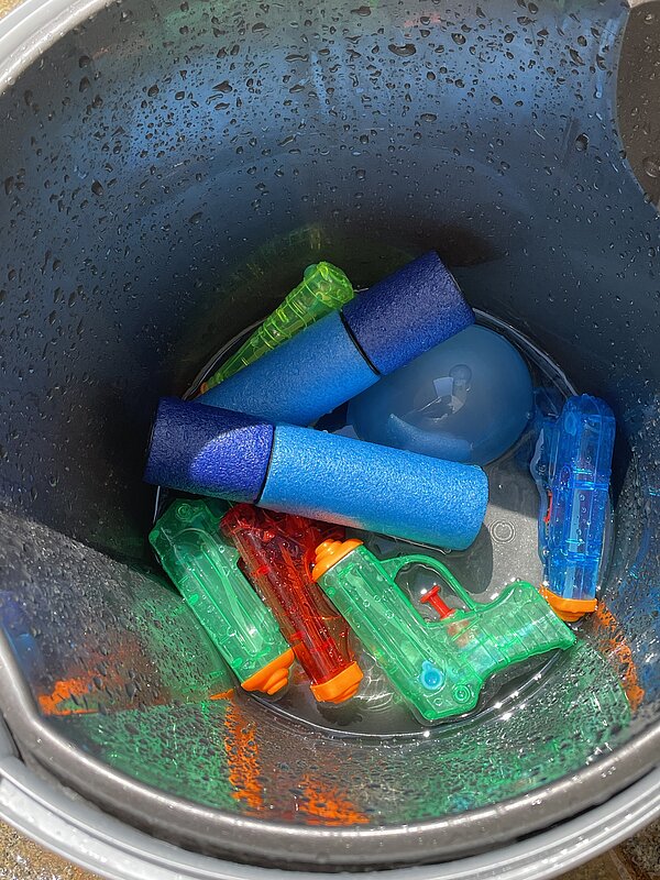 Nahaufnahme des schwarzen Eimers mit mehreren bunten Wasserpistolen, zwei blauen Schaumstoff-Wasserspritzen und einer gefüllten Wasserbombe.
