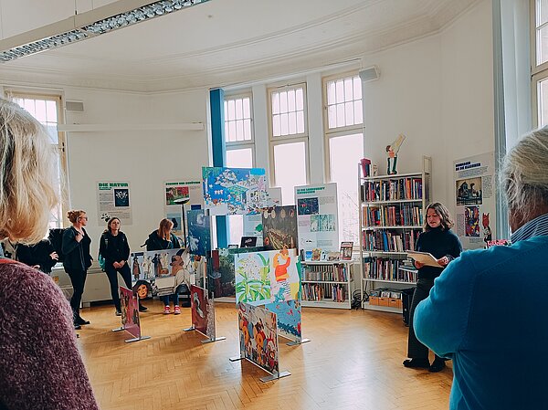 Buchmesse Leipzig: Wo Worte Bildung und Austausch fördern In einem Zimmer hellem Holzboden und Bücherschränken an den Wänden zwischen den großen Fenstern liest eine Frau etwas von einem Blatt ab. Außen herum stehen einige Zuhörer inmitten von aufgestellten Bildern, offenbar Illustrationen aus Büchern.