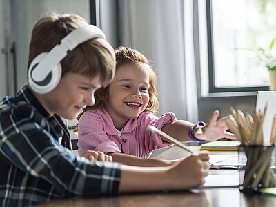 Zwei Kinder sitzen an einem Tisch und malen gemeinsam. Der Junge trägt Kopfhörer und konzentriert sich auf seine Zeichnung, während das Mädchen lächelnd neben ihm sitzt und ebenfalls malt. Auf dem Tisch liegen verschiedene Stifte und Malutensilien.