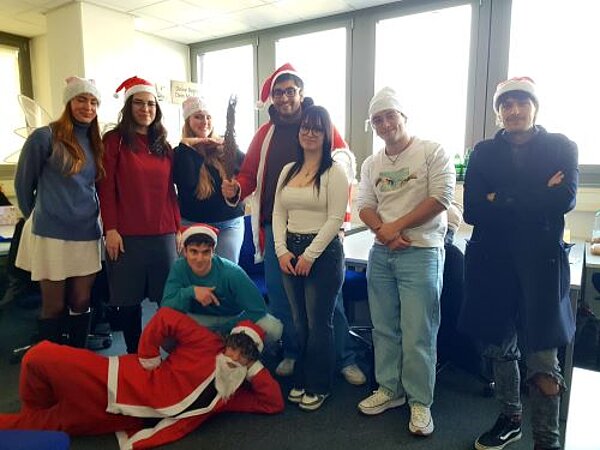 Eine Gruppe von acht Personen in einem Klassenraum vor großen Fenstern, die Weihnachtsmützen und festliche Kostüme tragen, posieren lächelnd für ein Foto. Einige stehen, während andere sitzen oder knien, und alle scheinen die festliche Atmosphäre zu genießen.