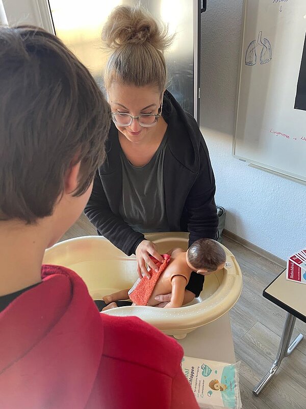 Eine erwachsene Person wäscht vorsichtig ein Baby in einer Babywanne. Eine weitere Person beobachtet den Vorgang. Im Hintergrund sind ein Flipchart mit Notizen und eine Packung Feuchttücher zu sehen.