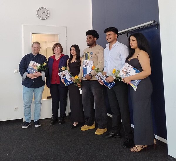 Sechs Personen stehen in einer Reihe in einem Raum mit blauer Wand und weißer Tür. Sie halten Blumensträuße und Zertifikate. Die Gruppe wirkt feierlich und stolz, möglicherweise bei einer offiziellen Veranstaltung. Eine Person trägt ein weißes, eine andere ein blaues Hemd, eine andere trägt ein beiges Longsleeve und eine weitere ein schwarzes Kleid.