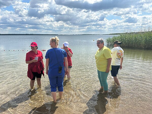 Ein Sinneskreis am Goitzsche-Strand: Lernen, Fühlen, Erleben! Fünf Personen stehen knöcheltief im Wasser am Seeufer. Sie unterhalten sich und genießen das Wetter, im Hintergrund Schilf.