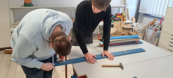 Ein junger Mann sägt ein Stück von einer blauen Holzlatte, die ein anderer junger Mann auf einem Tisch festhält.