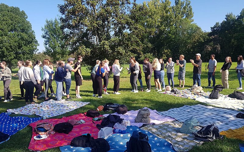 Eine Gruppe von Menschen stehen in einer Reihe auf einer Wiese in einem Park. Vor ihnen sind viele bunte Picknickdecken ausgebreitet. Der Himmel ist blau und die Sonne scheint.
