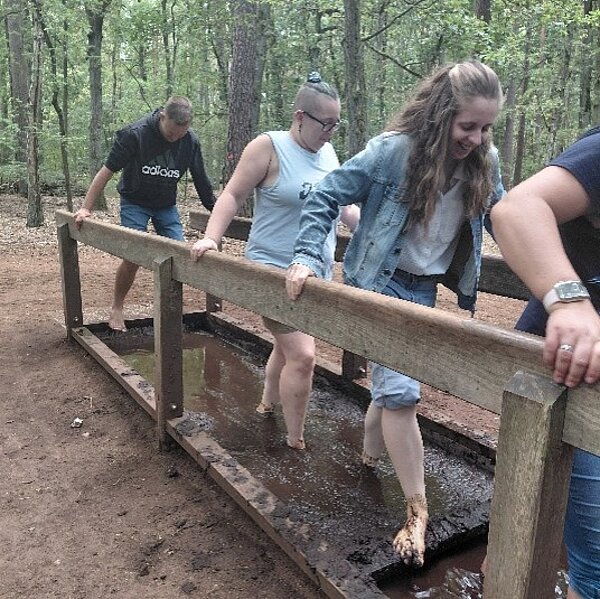 Vier Personen durchqueren barfuß ein flaches Becken mit Schlamm, während sie sich an einem Holzgeländer festhalten. Im Hintergrund sind Bäume und ein Waldweg zu sehen.