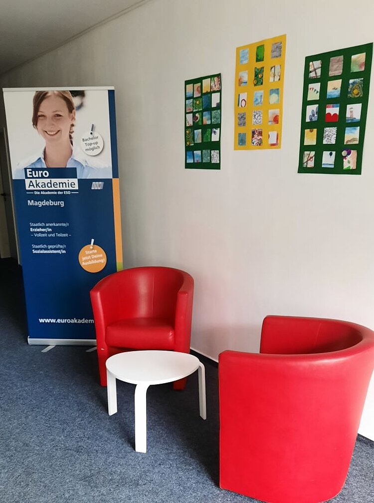 Ein Wartebereich mit zwei roten Sesseln und einem weißen Beistelltisch. An der Wand hängt ein Roll-up der Euro Akademie Magdeburg mit Informationen zu staatlich anerkannten Berufen wie Erzieher/in, Vollzeit- und Teilzeitausbildungen sowie sozialpädagogischen Weiterbildungen. Neben dem Roll-up sind bunte Collagen aus kleinen Bildern an der Wand befestigt.