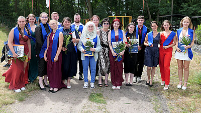 Auf einem Kiesweg vor Bäumen steht eine Gruppe junger Menschen in festlicher Kleidung. Sie tragen blaue Schärpen um die Schultern. Einige Menschen halten Blumen und Zeugnismappen in den Händen. In der Mitte steht ihre Lehrerin.
