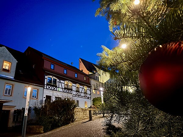 Nahaufnahme eines geschmückten Weihnachtsbaums mit Lichtern und roter Kugel im Vordergrund, historische Häuser und gepflasterte Straße im Hintergrund bei Nacht.