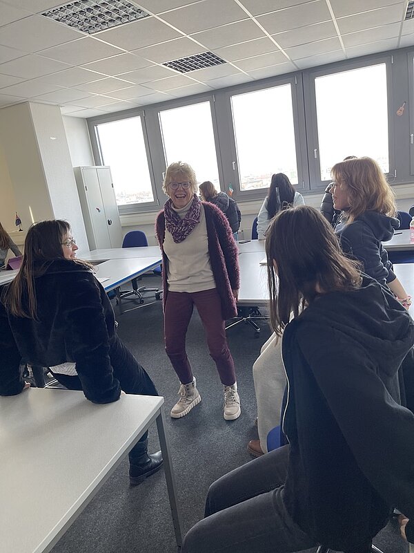 Eine Gruppe von Auszubildenden und Lehrkräften hat sich in einem Raum mit großen Fenstern versammelt. In der Mitte sitzt eine Person mit Brille, Schal und legerer Kleidung, die lächelt und sich mit den anderen unterhält. Der Raum ist mit Tischen und Stühlen ausgestattet, und die Anwesenden sitzen oder lehnen sich an die Möbel und unterhalten sich angeregt. Die Atmosphäre wirkt ungezwungen und entspannt.