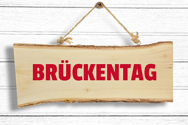 Wichtiger Hinweis in eigener Sache! Ein Holzschild mit der Aufschrift "BRÜCKENTAG" hängt an einer weißen Holzwand.