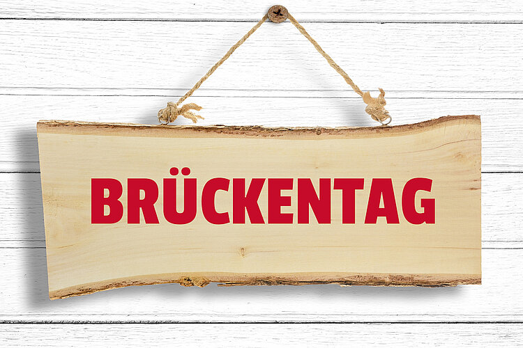 Wichtiger Hinweis in eigener Sache! Ein Holzschild mit der Aufschrift "BRÜCKENTAG" hängt an einer weißen Holzwand.