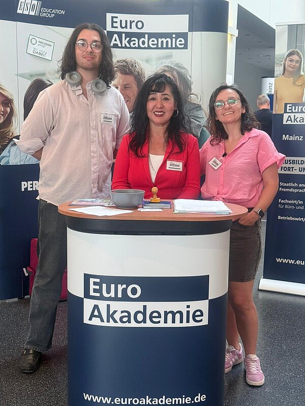 Euro Akademie Mainz auf BIM Rheinhessen Drei Personen – zwei Lehrkräfte und ein Schüler – stehen lächelnd hinter dem Informationsstand der Euro Akademie Mainz. Auf dem Tisch liegen Broschüren und ein Stempel, während im Hintergrund ein Roll-Up mit dem Logo der Euro Akademie zu sehen ist.