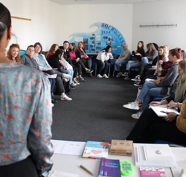 Theorie meets Praxis Blick aus der Perspektive einer Referentin auf die Klasse, die links und rechts in zwei Reihen auf Stühlen sitzen. Die Referentin ist von hinten halb zu sehen. Sie steht vor einem Tisch, auf dem Bücher, Dokumente und Unterrichtsmaterialien liegen.