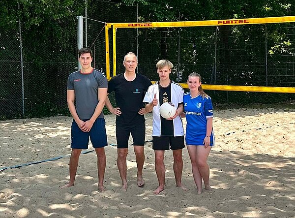 Beachvolleyball mitten in Hannover Vier Personen, darunter drei Schüler*innen und ein Lehrer der Euro Akademie Hannover, stehen barfuß auf einem Beachvolleyballfeld vor dem Netz. Sie lächeln in die Kamera, eine Person hält einen Volleyball in der Hand.