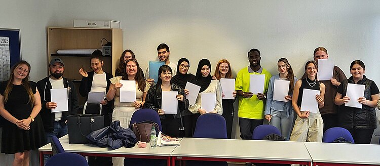 Eine Gruppe junger Erwachsener steht in einem Klassenraum und hält stolz ihre Zeugnisse oder Teilnahmebescheinigungen in den Händen. Alle lächeln in die Kamera, im Hintergrund sind ein Regal, ein Kalender und Tische mit Taschen zu sehen.