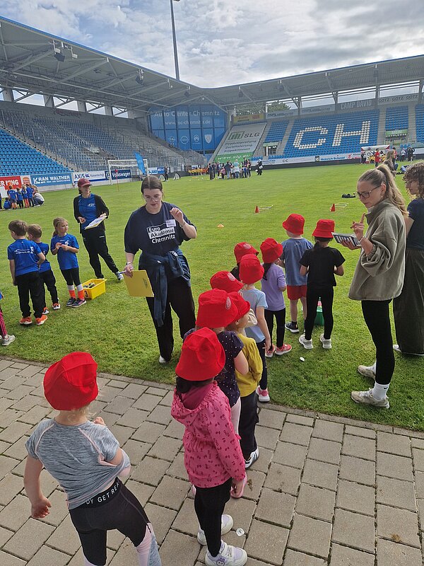Kinder mit roten Sonnenhüten stehen mit Betreuungspersonen und Mitarbeitenden der Euro Akademie Chemnitz am Spielfeldrand eines Stadions und warten auf ihre nächste Aktivität.
