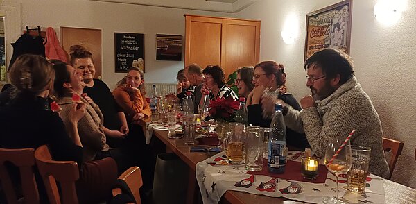 Lange Tafel mit lachenden Gästen in einer Gaststube; Kerzen, Poinsettie und Getränke sorgen für stimmungsvolle Weihnachtsatmosphäre.