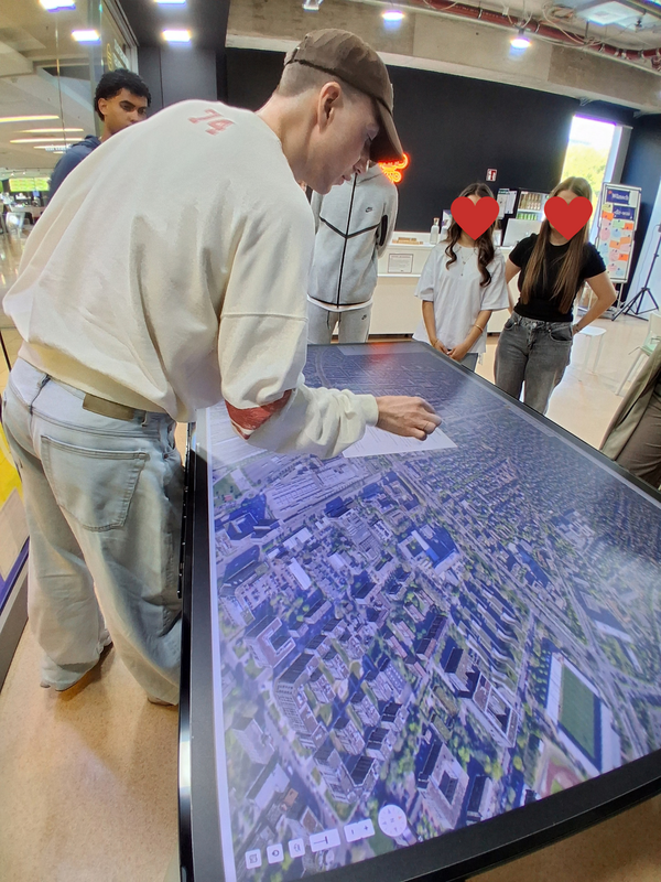 Ein junger Mann berührt einen großen horizontalen Bildschirm mit einem Stadtplan. Daneben stehen weitere Personen.