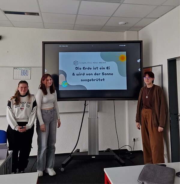 Drei Schülerinnen präsentieren eine Folie mit dem Titel „Die Erde ist ein Ei & wird von der Sonne ausgebrütet“. Die Folie ist bunt illustriert und humorvoll gestaltet.