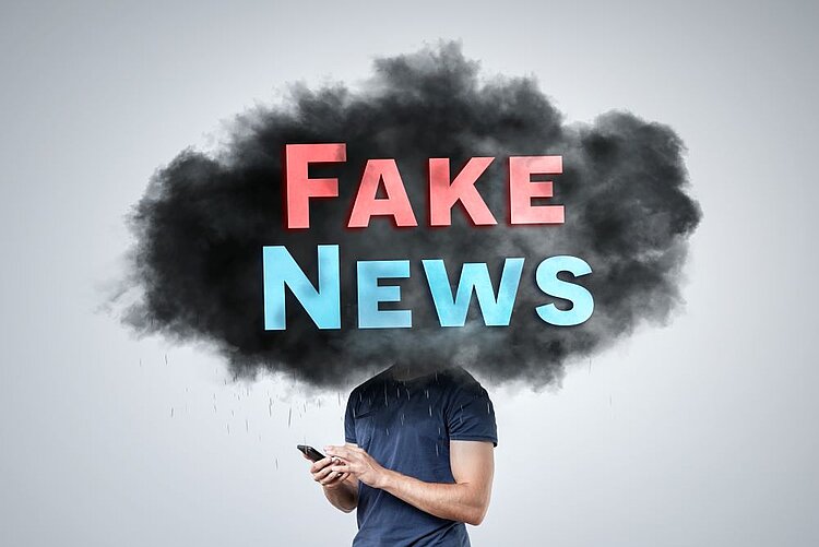 Demokratieprojekt Ein Mensch steht unter einer dunklen Wolke, die die Worte "Fake News" in leuchtenden Farben zeigt, während er auf ein Smartphone schaut. Das Bild symbolisiert die allgegenwärtige Bedrohung durch Fehlinformationen in der modernen digitalen Welt.