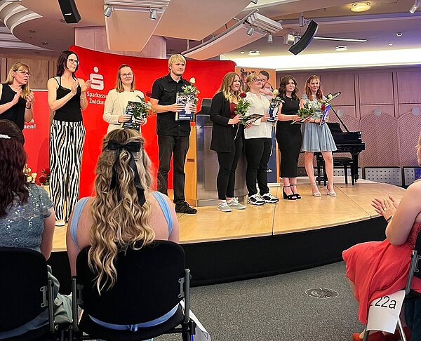 Feierlicher Abschied: Exmatrikulation der Absolvent*innen 2025 Eine Gruppe von Personen steht auf einer Bühne und hält Blumen und Zertifikate, zwei klatschen. Sie sind elegant gekleidet und lächeln. Im Hintergrund ist ein rotes Banner und ein Klavier zu sehen.