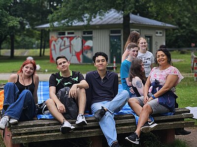 Schülerinnen und Schüler sowie eine Lehrerin sitzen auf einer hölzernen Bank mit Picknickdecke und posieren für ein Foto.