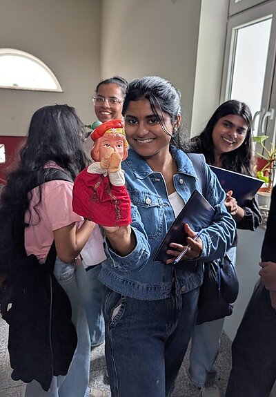 Eine Auszubildende der Euro Akademie mit langen dunklen Haaren trägt eine blaue Jeansjacke und hält eine Handpuppe mit rotem Gewand und roter Mütze in die Kamera. Im Hintergrund stehen weitere Personen, von denen eine ein rosafarbenes Oberteil trägt. Die Szene spielt in einem hellen Raum mit großen Fenstern und einer Topfpflanze auf der Fensterbank.