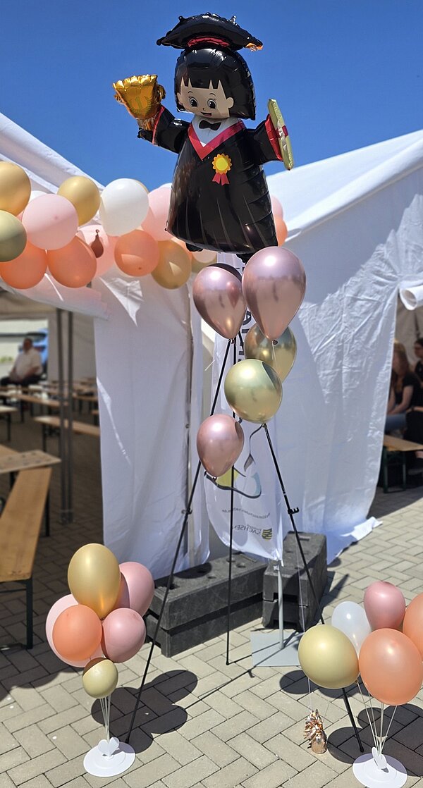Blick auf Holzbänke und Tische, die unter weißen Leinentüchern stehen, davor Luftballons und eine Ballon-Figur mit Absolventenhut als Dekoration.