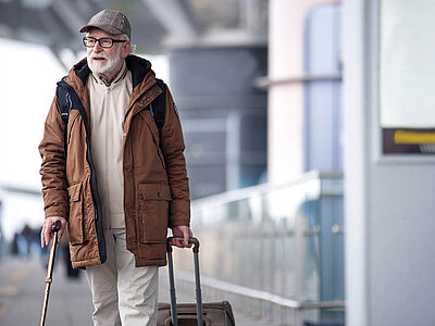 Ein älterer Mann mit weißem Bart, Brille und einem braunen Hut läuft mit einem Rollkoffer durch einen Flughafen.