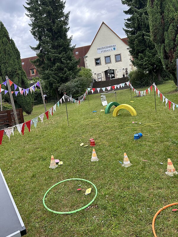 Großer Spielparcours auf einer Wiese vor dem Gebäude des Freizeitzentrums Pößneck. Der Parcours ist mit bunten Wimpelketten, Hula-Hoop-Reifen, kleinen Hindernissen und weiteren Spielmaterialien aufgebaut.