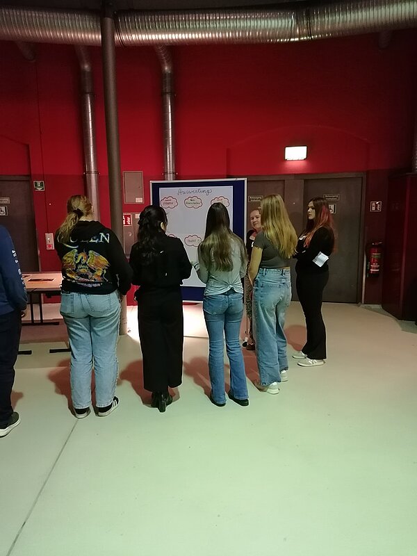 Schülerinnen und Schüler stehen vor den erarbeiteten Inhalten, die verschriftlicht und anschaulich festgehalten wurden, vor einem Flipchart.