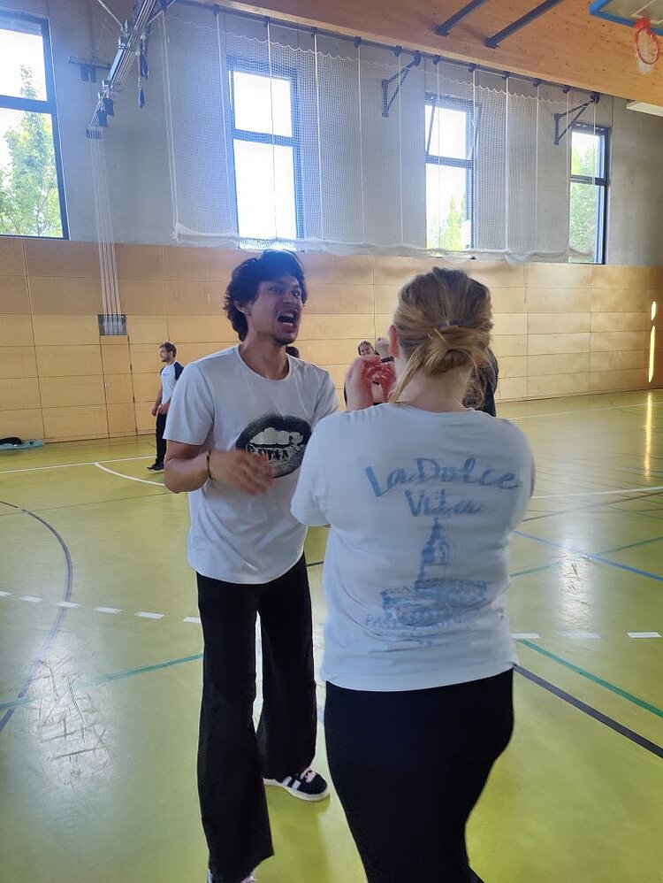 Ein Mann und eine Frau in einem hellen Sportraum mit großen Fenstern und Holzpaneelen. Der Mann, der ein weißes T-Shirt und schwarze Hosen trägt, steht einer Frau gegenüber, die ein weißes T-Shirt mit der Aufschrift 'La Dolce Vita' und einem Rückendruck eines Gebäudes trägt. Der Mann hat seinen Mund weit geöffnet und scheint die Frau anzuschreien.