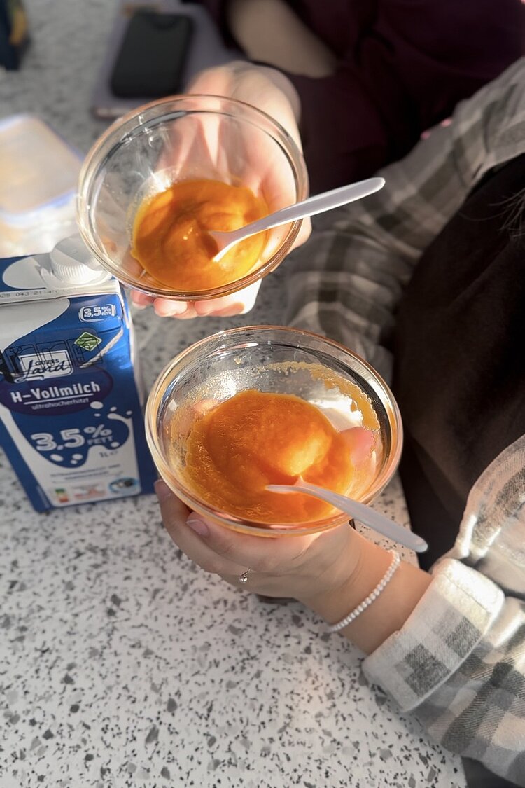 „Ein Löffel für Mama…“ Hände von Auszubindenden halten zwei Glasschüsseln mit Löffen und selbstgemachtem Brei in die Kamera, daneben steht ein Tetrapak Milch.