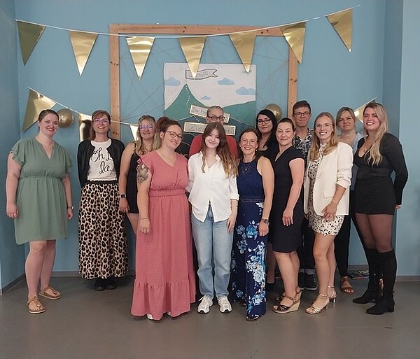 Herzlichen Glückwunsch! Absolventinnen und Absolventen posieren bei Abschlussfeier für Erinnerungsfoto. Sie tragen sommerliche und festliche Kleidung, stehen vor einer pastellblauen Wand, mit Girlanden und Wimpeln aus Gold im Hintergrund.