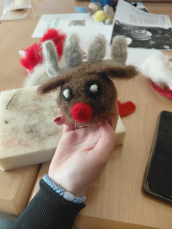Kleine gefilzte Rentierfigur mit roter Nase und grauen Geweihen wird von einer Hand über einer Filzunterlage gehalten.