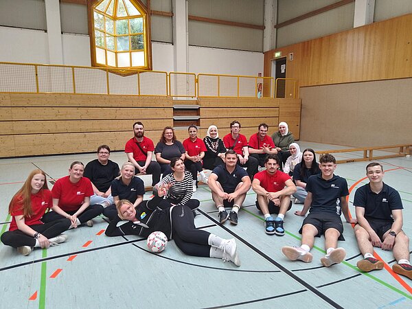 Eine Gruppe von Teilnehmenden sitzt gemeinsam in einer Sporthalle auf dem Boden und posiert für ein Gruppenfoto; im Vordergrund liegt ein Fußball.