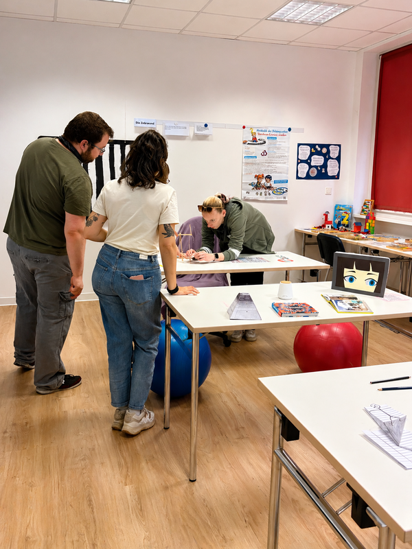 Mehrere Personen arbeiten gemeinsam an einer Station mit verschiedenen Lern- und Experimentiermaterialien in einem Unterrichtsraum.