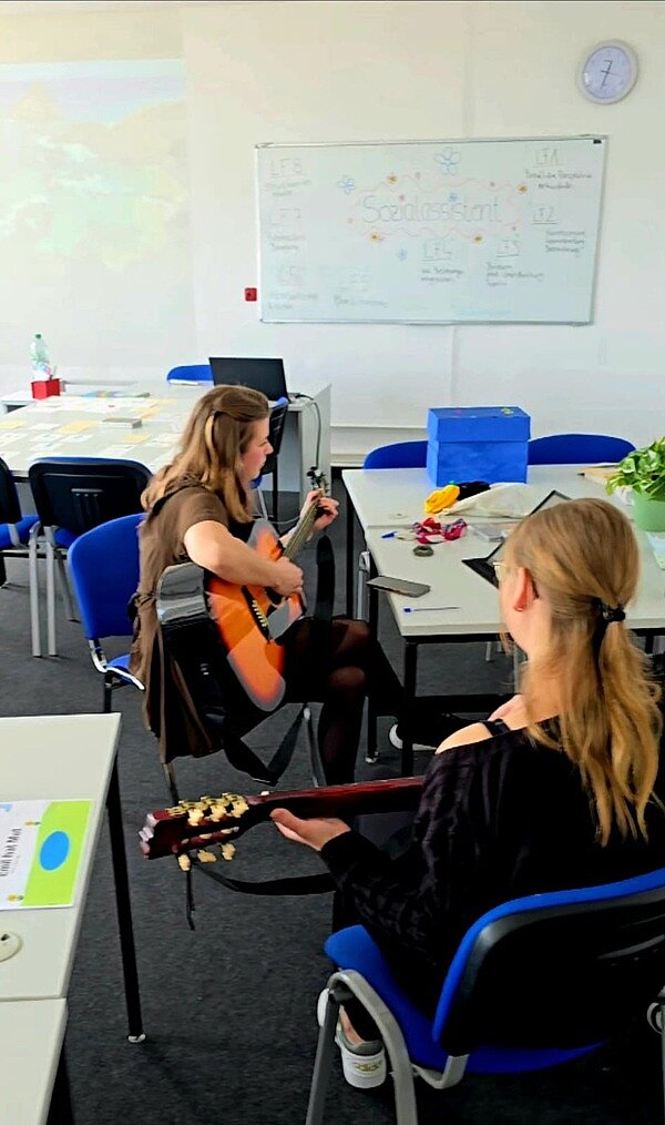 Zwei weibliche Teilnehmende sitzen in einem Klassenraum und beide spielen Akustikgitarre.